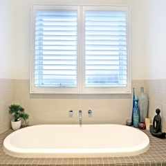White Bathroom Plantation Shutters with 2 panel doors mounted on window above bath tub in semi open position